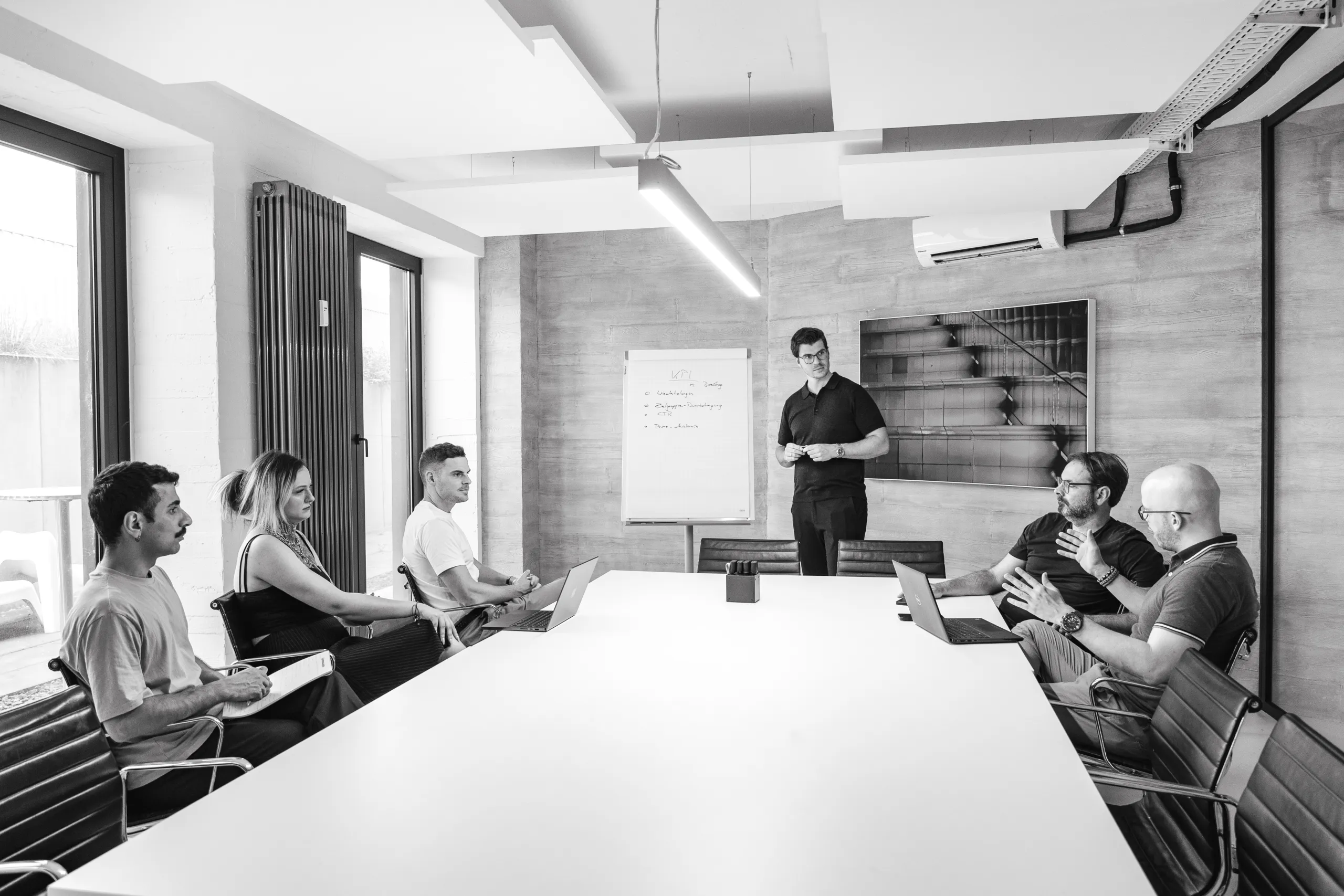 Sechs Mitarbeitende von GRAPEVINE in einer Besprechung am Konferenztisch, einer steht vor einem Flipchart mit KPIs.